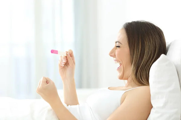 在家里的床上检查阳性妊娠试验的兴奋妇女的侧面图 — 图库照片