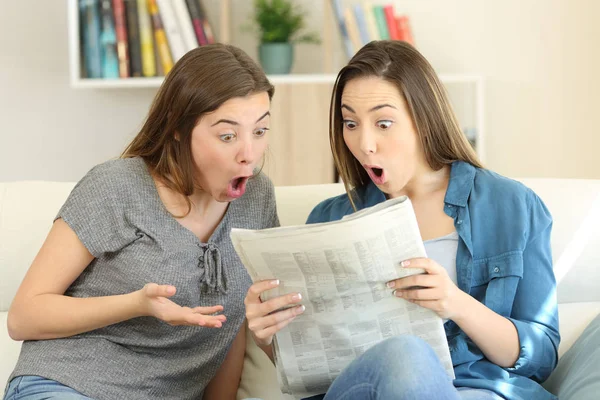 Amazed friends reading newspaper news sitting on a couch in the living room at home