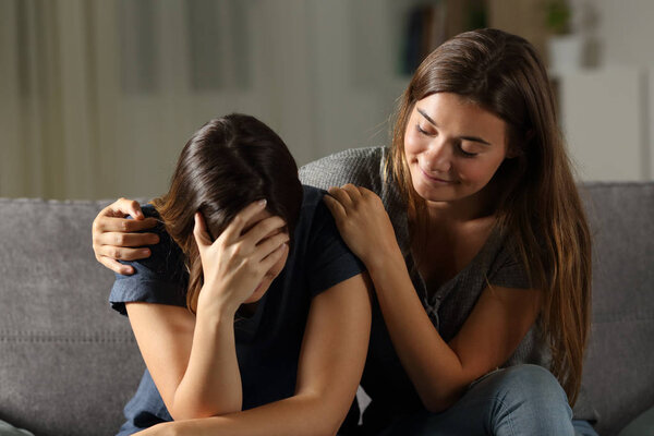 Bad hypocrite friend comforting a sad girl sitting on a couch in the living room at home 