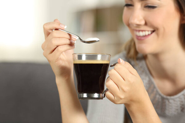 Close up of a happy woman hand throwing sugar into coffee with a spoon sitting on a couch in the living room at home