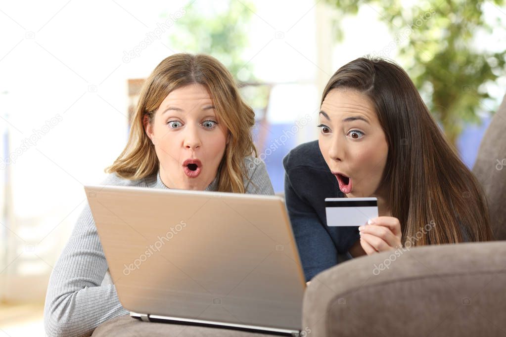 Front view portrait of an amazed friends buying online together sitting on a couch in the living room at home