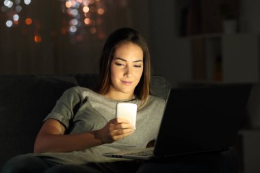 Oturma odasında evde bir kanepede oturan gecede bir dizüstü bilgisayar ve bir telefon kullanan rahat kız