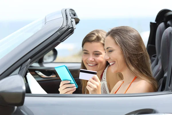 Side view portrait of two tourists paying online with a smart phone and credit card inside a rental car on vacation
