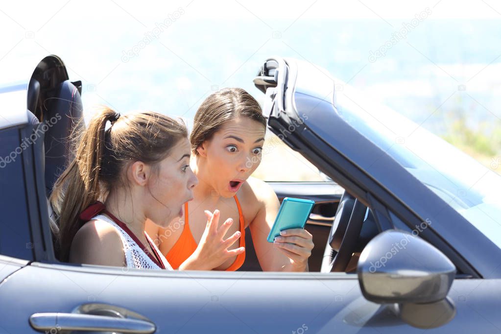 Two surprised tourists inside a convertible car reading good news in a phone on vacation
