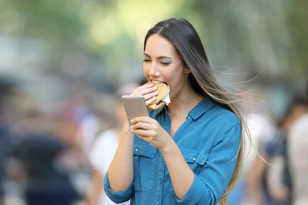 Szczęśliwa Kobieta Jedzenie Burger Gospodarstwo Inteligentny Telefon Ulicy — Zdjęcie stockowe