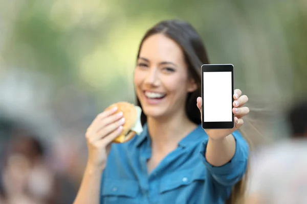 女人拿着一个汉堡包显示一个空白的电话屏幕模拟在街上 — 图库照片