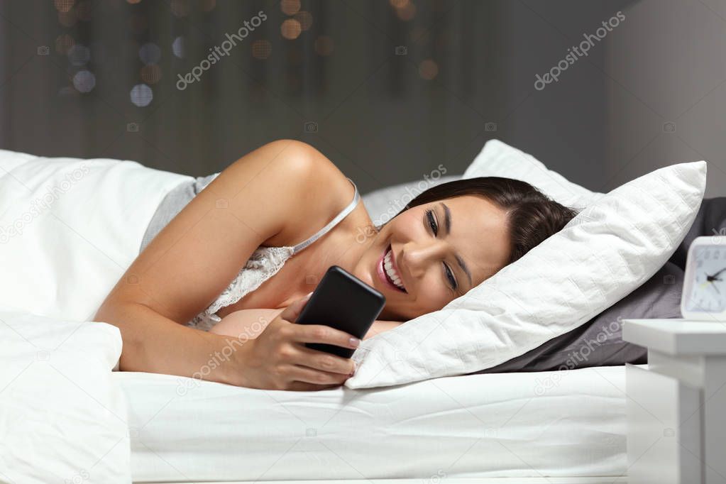 Happy woman checking smart phone in the night lying on the bed at home