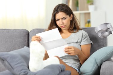 Worried disabled woman reading a letter sitting on a couch in the living room at home clipart