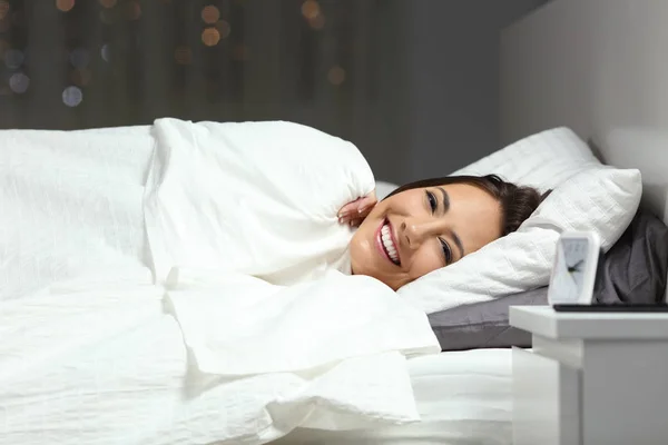 Happy woman covering with a blanket in the bed looking at camera in the night at home