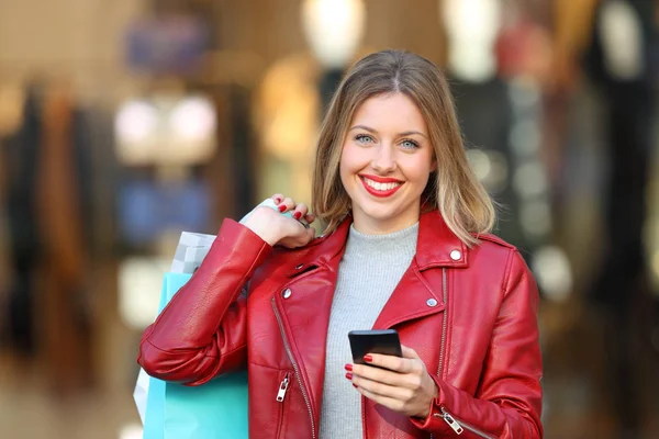 快乐的购物者摆在街上拿着一个智能手机和购物袋的背景下的店面 — 图库照片