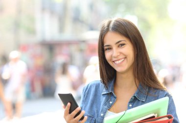 Mutlu öğrenci kamera tutan bir akıllı telefon ve klasörleri sokakta bakarak poz
