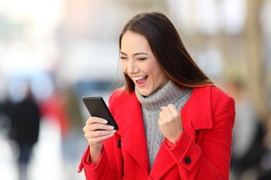 Portrait of an excited woman in red reading good news in a smart phone in winter on the street clipart