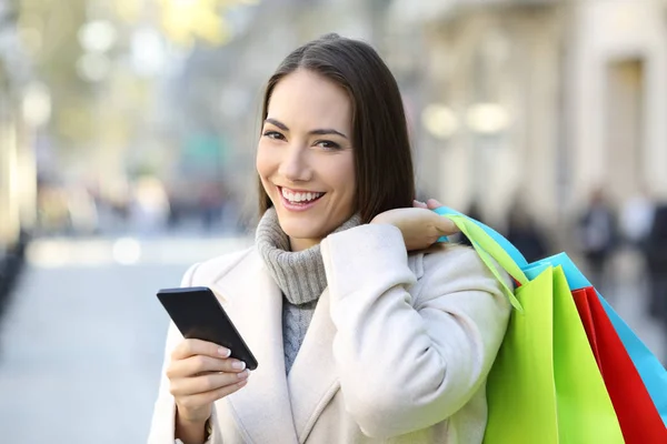 快乐购物者手持智能手机和五颜六色的购物袋在冬天在街上 — 图库照片