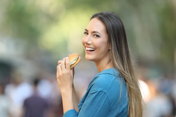 通りでカメラ目線ハンバーガーを食べて幸せな女 — ストック写真