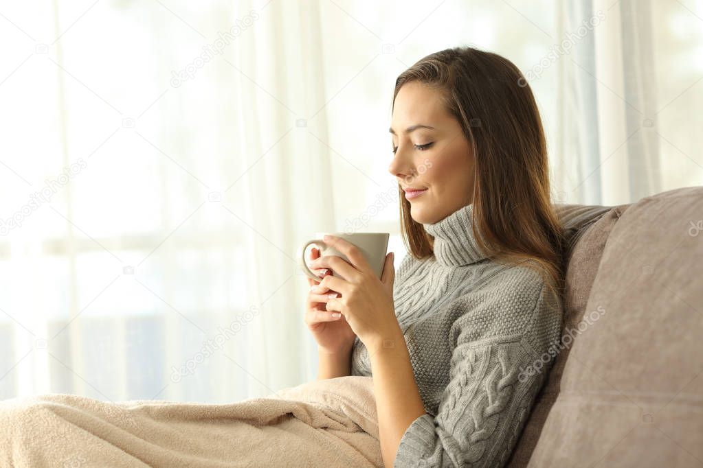 Pensive lady holding a cup of coffee in winter sitting on a couch in the living room at home