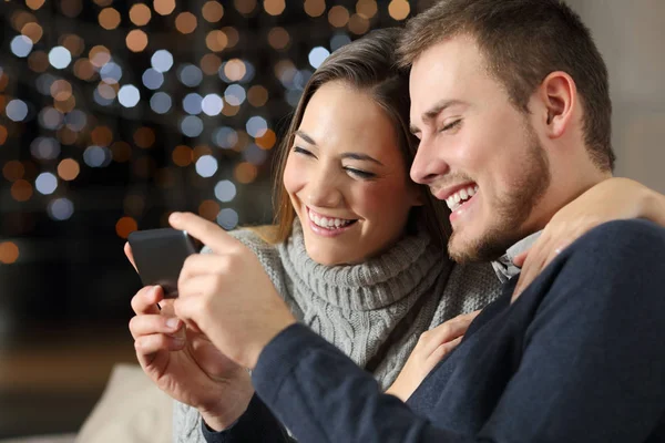 Couple watching videos in a phone in the night sitting on a couch in the living room at home