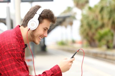 Yan görünümü ile telefon ve kulaklık bir tren istasyonunda bekleyen müzik dinlerken mutlu bir adam portresi