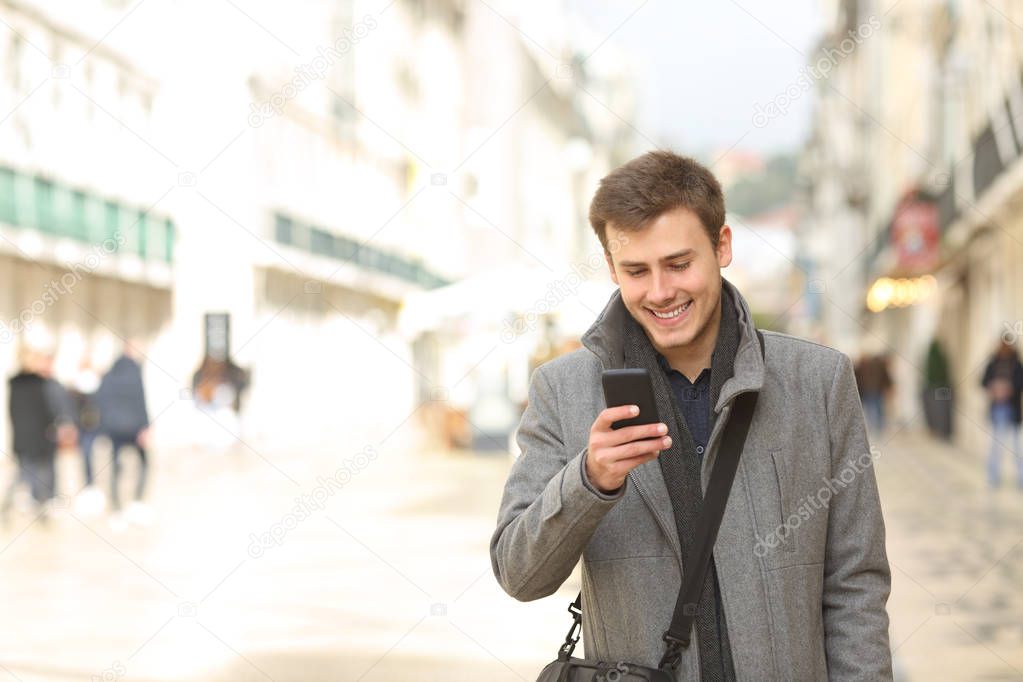 Front view portrait of a happy man walking using a smart phone in winter in the street