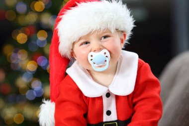 Bebek evde oturma odasında kanepede Noel kameraya bakıyor ağzında emzik ile Santa kılık giymiş