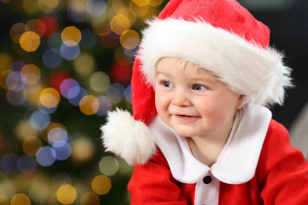 一个婴儿的肖像穿着圣诞老人伪装看着在家里的沙发上与树和背景灯的一侧 — 图库照片
