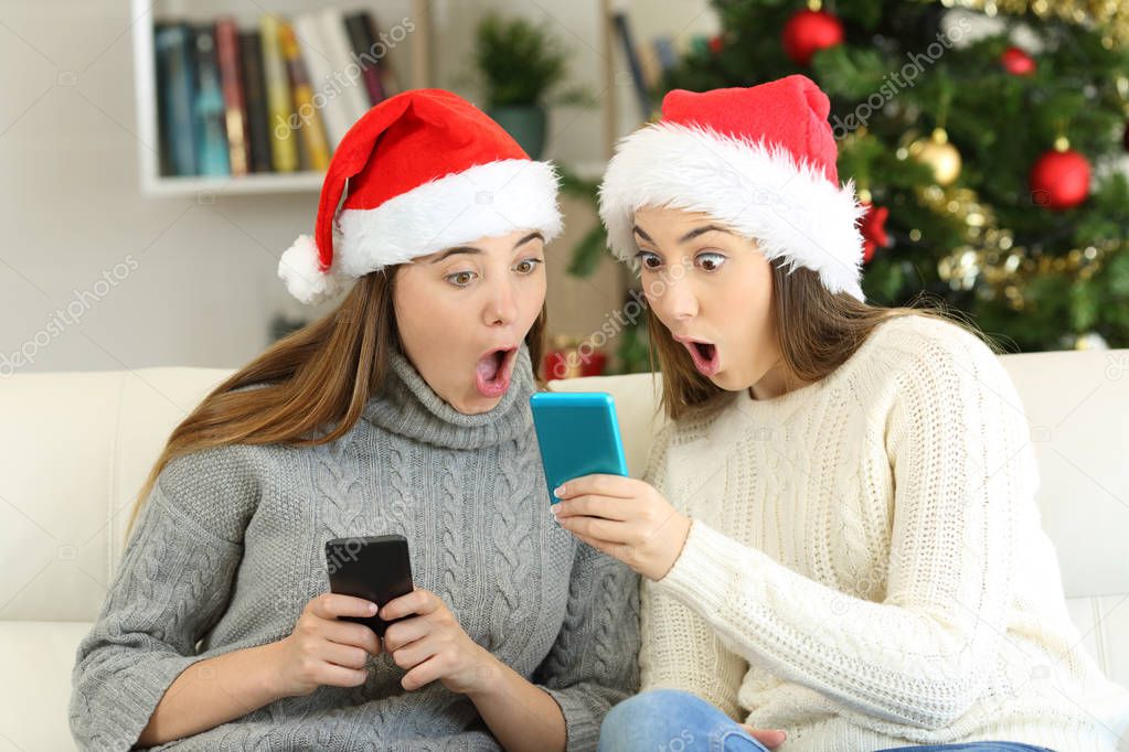 Two amazed friends finding online offers in christmas sitting on a couch in the living room at home