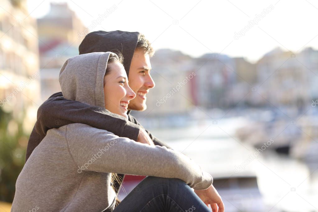 Side view portrait of a happy couple of teenagers in love contemplating a coast town on vacation