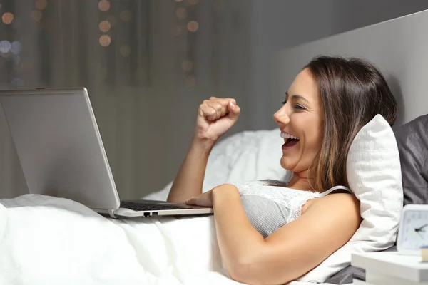 一个兴奋的妇女的侧面视图肖像庆祝好在线新闻在床上晚上在家 — 图库照片
