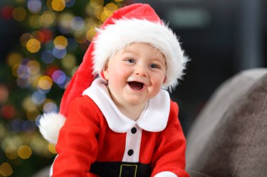 Neşeli bebek Noel Baba olarak evde bir kanepede Noel içinde bir ağaç arka planda giyinmiş