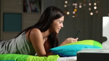 Mutlu bir gencin gece yatağında uzanmış akıllı bir telefonla medyayı izlemesinin yan görüntüsü.