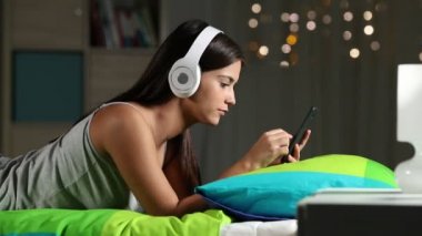 Yan görünümü gece evde bir yatakta telefon ve kulaklık ile müzik dinlemek mutlu bir genç portresi