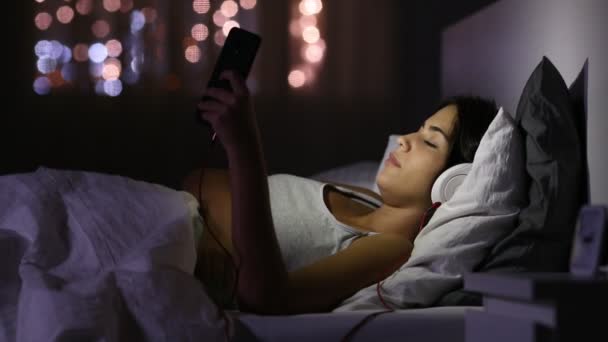 歌と夜に自宅のベッドで携帯電話で音楽を聴く幸せなティーンの側面図 — ストック動画