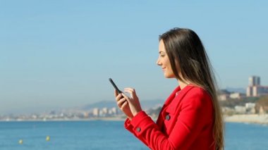 Yan görünümü kışın sahilde bir akıllı telefon kullanarak heyecanlı bir kadın portresi. Ağır çekim