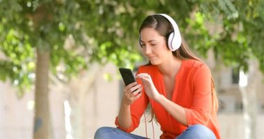 Turuncu bir parkta bir bankta oturmuş bir telefondan müzik dinlerken kulaklık giyen mutlu kadın