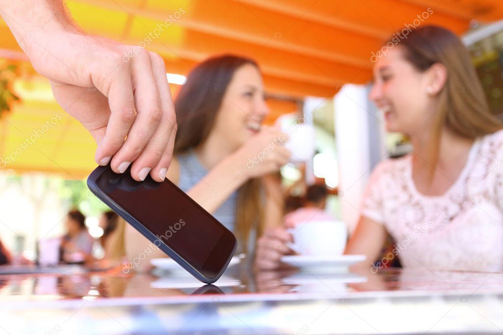 Distracted friends talking in a bar and close up of a thief hand stealing phone on foreground