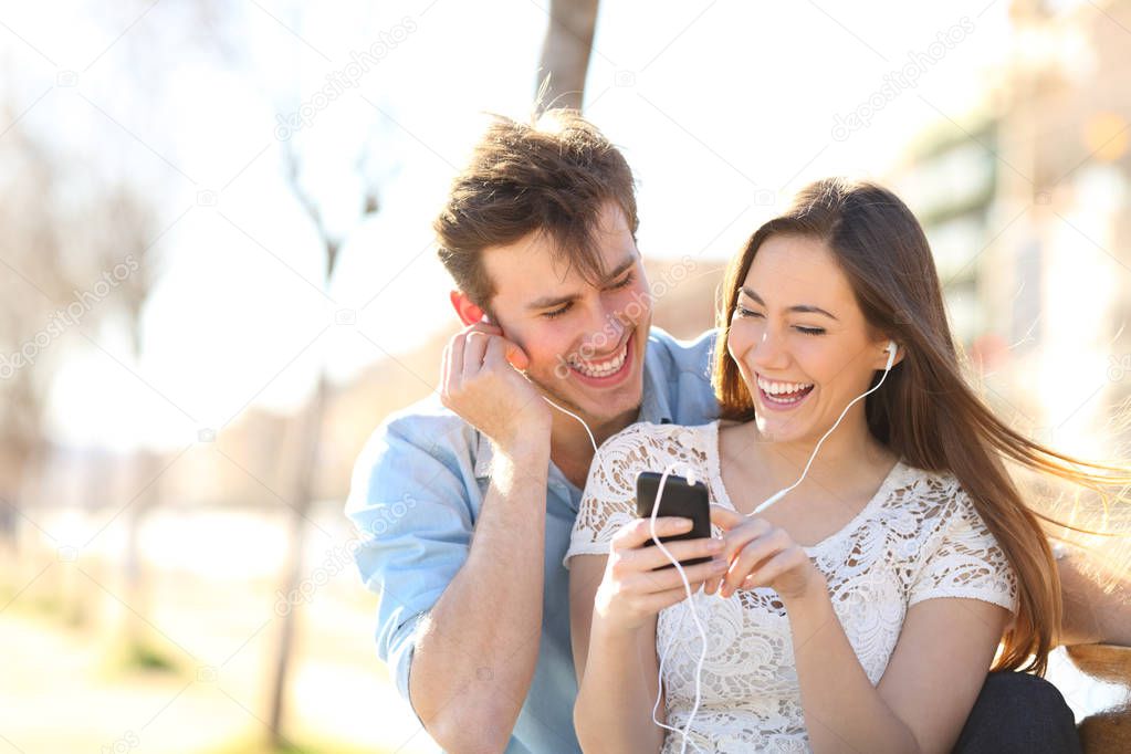 Joyful couple listening to music from a smartphone sitting on a bench in a pak