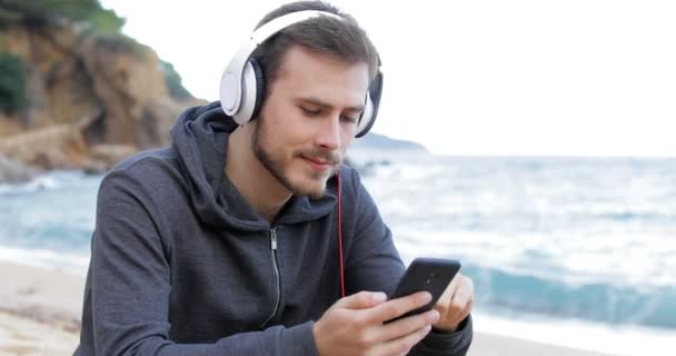 严重的十几岁的男孩下载和听音乐从一个智能手机在海滩上 — 图库视频影像