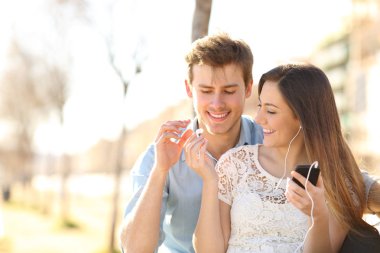 Mutlu çifte bir parkta bir bankta oturmuş bir akıllı telefondan müzik dinlemek için kulaklık paylaşımı