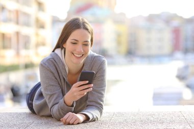Mutlu genç kız akıllı telefon içeriğini kontrol ediyor. Bir sahil kasabasının kaldırımında yatıyor.