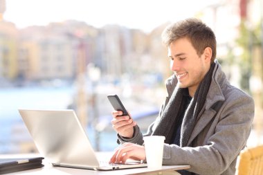Bir sahil kasabasının bar terasında akıllı telefon ve laptop kullanan mutlu girişimci.