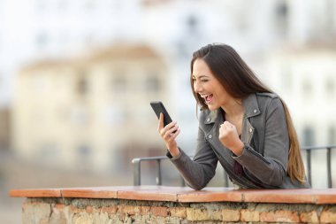 Excited woman checking smart phone in a balcony with a town in the background clipart