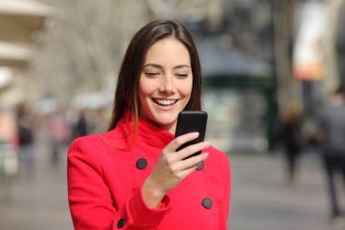 Kırmızı ceket giyen mutlu kadın kışın sokakta akıllı telefon kullanarak yürüyor.