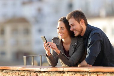 Mutlu çift arka planda bir kasaba ile günbatımında bir balkonda akıllı telefon kullanma