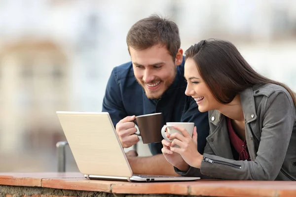 愉快的夫妇检查笔记本电脑在线内容拿着咖啡杯在阳台上与一个小镇的背景 — 图库照片