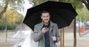 Bir şemsiye yağmur altında tutan kameraya doğru yürümek telefon kontrol mutlu bir adam portresi önden görünüm