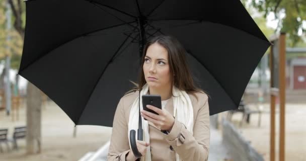 冬の雨の下でスマート フォンで天気予報アプリをチェック 欲求不満な女の正面肖像画 — ストック動画