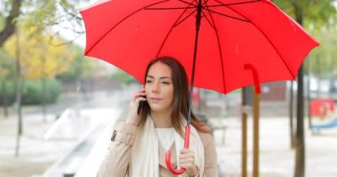 Mutlu bir kadın telefon üzerinde yürüyen bir parkta yağmur altında kırmızı şemsiye tutan söz portresi önden görünüm
