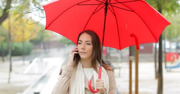 電話歩行公園で雨の中赤い傘を保持に関する話幸せな女の正面肖像画 — ストック動画