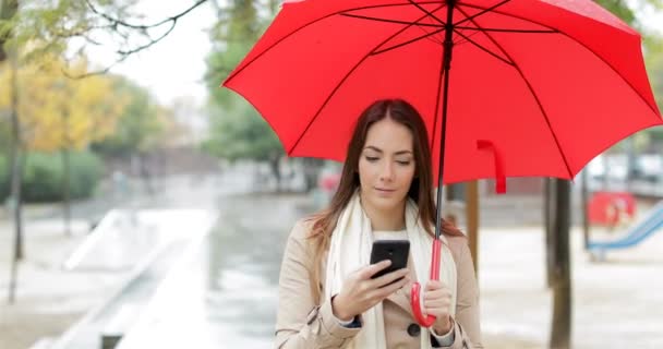 スマート フォンは 雨の中赤い傘を持って歩いてチェック深刻な女性の正面肖像 — ストック動画