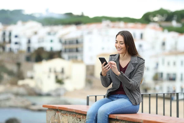 Happy woman sitting on a ledge using a smart phone in a coast town