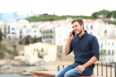 Mutlu adam akıllı telefonda konuşuyor. Sahil kasabasında tatilde bir çıkıntıda oturuyor.